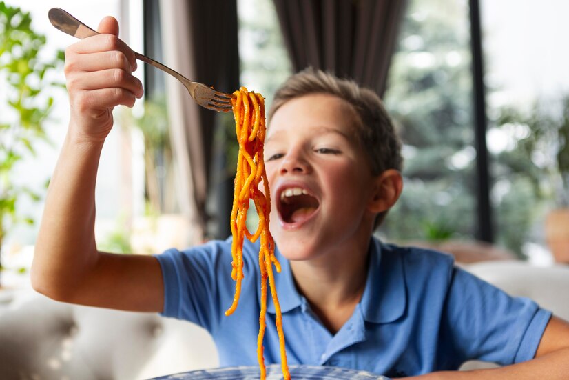 Young boy about 12 years old in a blue shirt eating spaghetti with a big smile. 